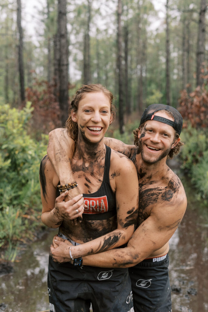 couple smile at the camera, covered in mud in the middle of the woods during their dirt bike engagement shoot