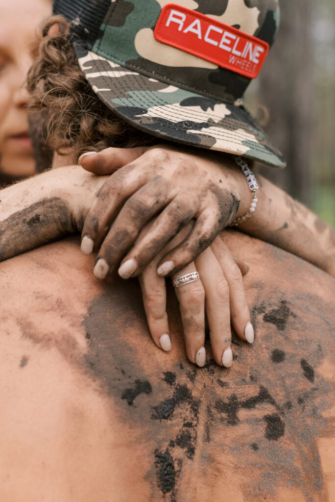 close up of muddy fingers as a woman holds her boyfriend close, he wears a camo hat backwards